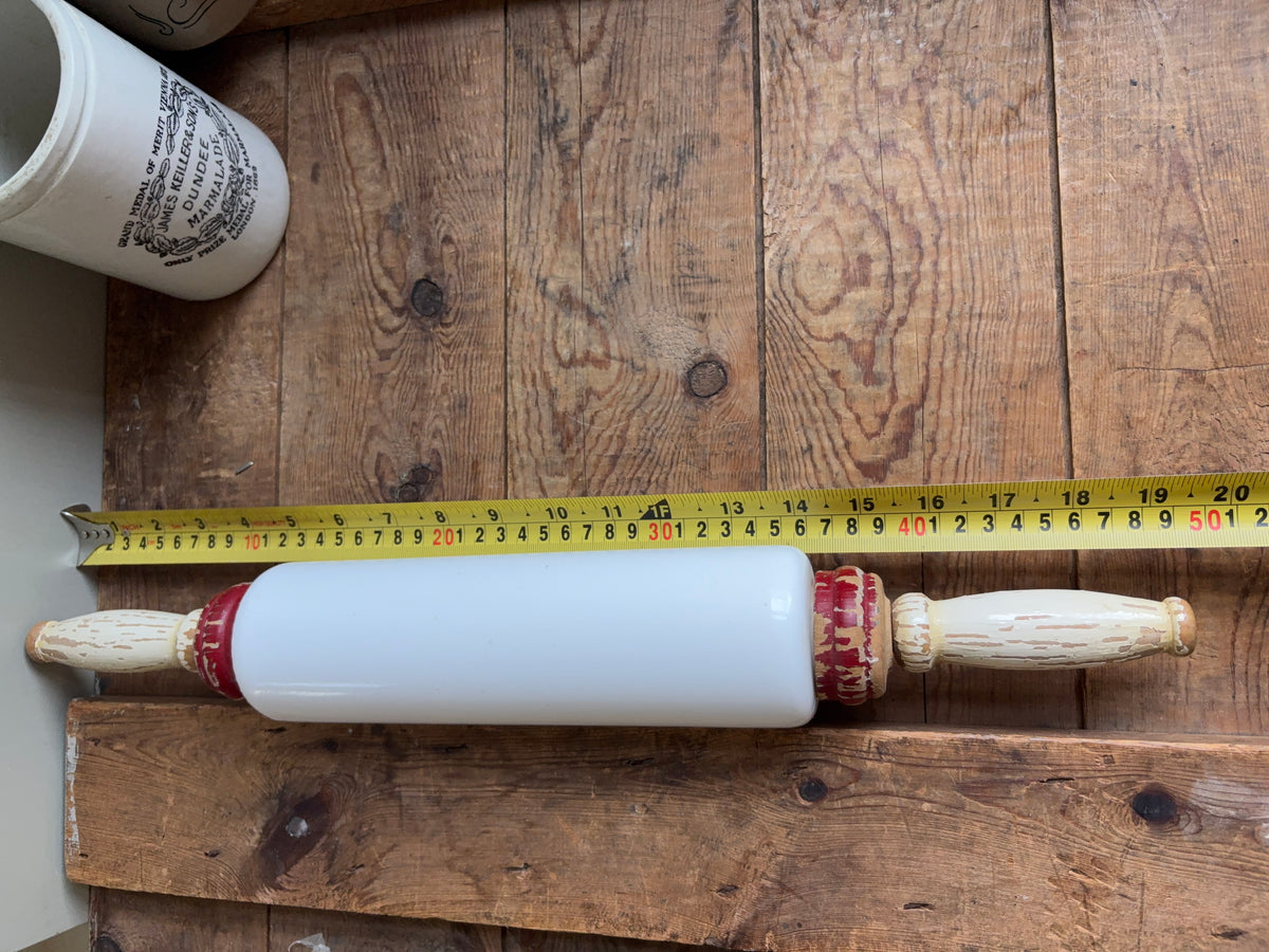 A white ceramic rolling pin with red painted accents and distressed cream handles, shown on a rustic wooden surface with tape measure.